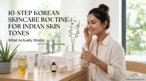 A smiling Indian woman with glowing skin applying a hydrating essence in a bright, modern bathroom, demonstrating a customized 10-step Korean skincare routine for Indian skin tones, featuring a lineup of minimalist K-Beauty products and thefashioncounty.com branding.